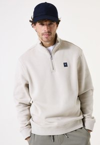 Sudadera beige con cremallera y cuello alto, de tejido texturizado y parche de logo en el pecho; combinado con una gorra azul marino.