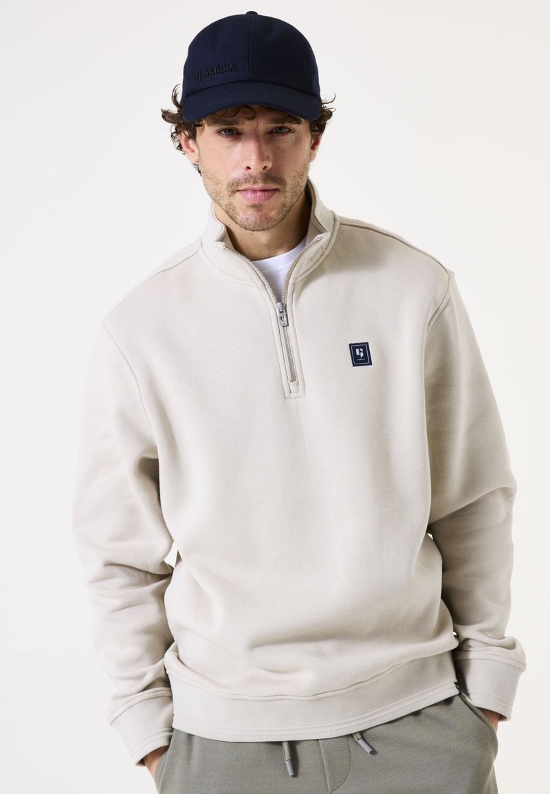 Sudadera beige con cremallera y cuello alto, de tejido texturizado y parche de logo en el pecho; combinado con una gorra azul marino.