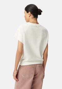 Sudadera blanca de manga corta con un corte relajado, que presenta un dobladillo de canalé, una textura suave y detalles en la parte posterior. Combinada con pantalones rosas claros.