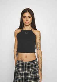 Black cropped tank top with a scalloped hem and "Tommy" logo. The model wears a plaid skirt with pleats and visible tattoos on her arms.