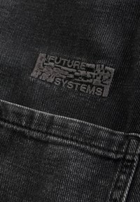 Svart denimtyg med en subtil struktur, med en präglad logotyp som säger "FUTURE SYSTEMS" på vänstra fickområdet.