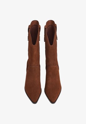 Par de botas de tornozelo em camurça castanha com bicos pontudos, saltos baixos, alças laterais e forro interior suave, mostradas de cima sobre fundo branco.