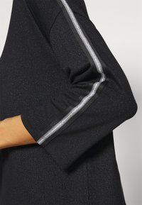 Czarny top wykonany z teksturowanego, błyszczącego materiału. Posiada rękawy o długości do łokcia z srebrnym paskiem wzdłuż szwów.