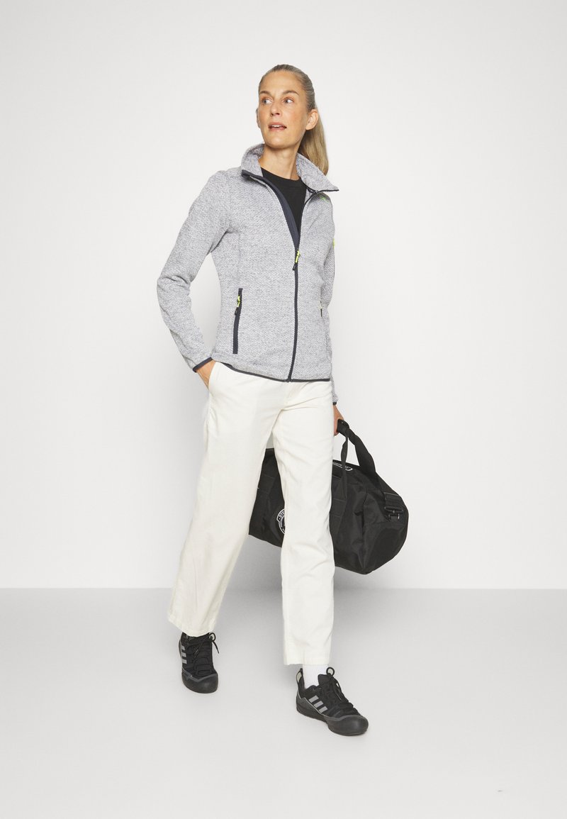 Giacca in pile grigia con tasche con zip, colletto alto, abbinata a pantaloni chiari e sneakers nere, mentre tiene un borsone nero.