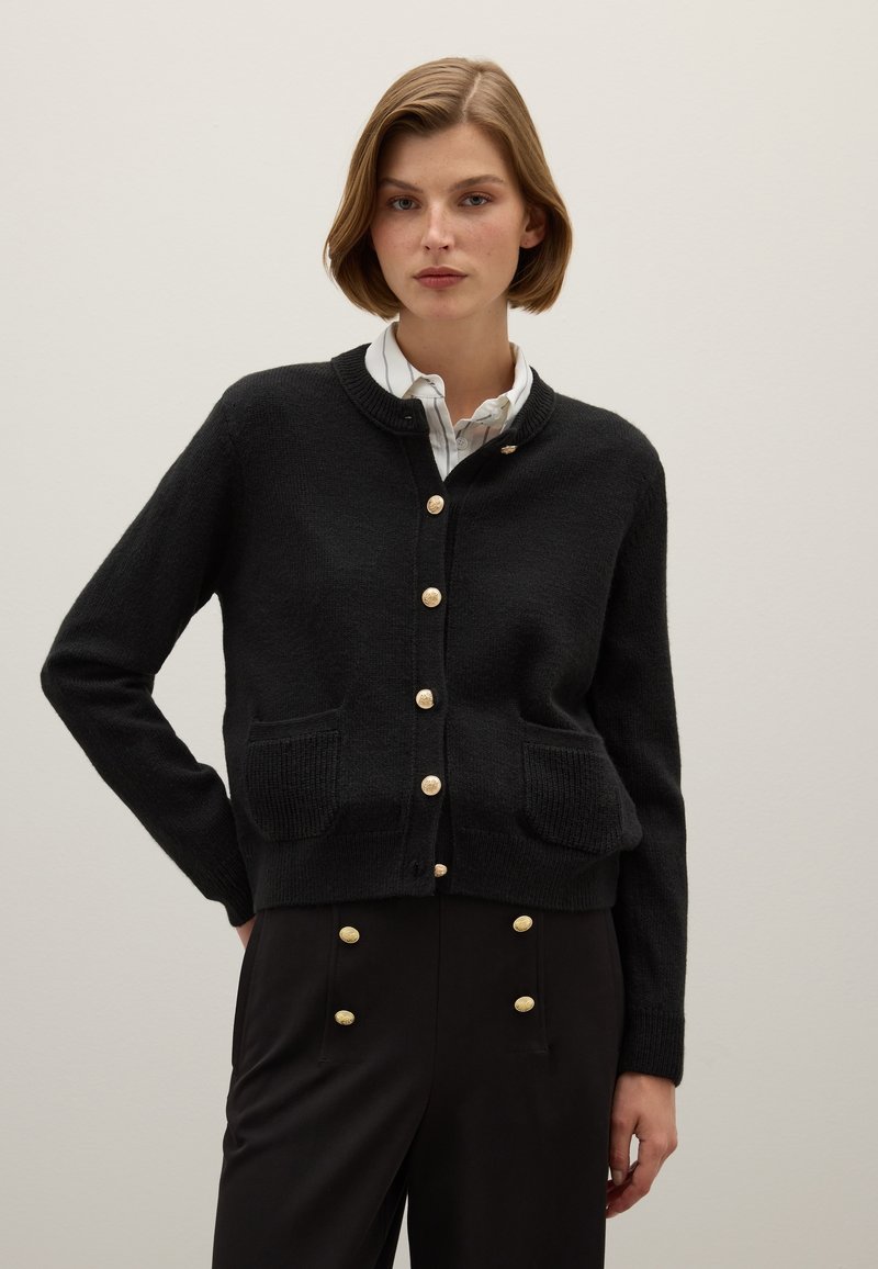 Cardigan noir en tissu texturé, avec un col rond, des boutons dorés et deux poches avant. Porté sur une chemise blanche à col.