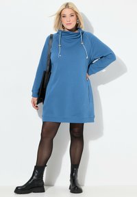 Blaues Sweatkleid mit einem Kapuzenkragen, Zugbandschmuck, langen Ärmeln und Seitentaschen, kombiniert mit schwarzen Knöchelstiefeln und Strumpfhosen.