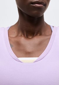 Κοντινό πλάνο του λαιμού και του πάνω μέρους του στήθους ενός ατόμου που φοράει μωβ μπλούζα με V λαιμόκοψη πάνω από λευκό ένδυμα, με τα χείλη και μέρος του κάτω προσώπου ορατά.