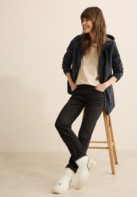 Marinblå jacka med huva, beige tröja, svarta jeans och vita högskurna sneakers. Funktioner inkluderar elastiska ärmslut och en avslappnad design.