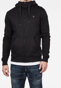 Svart zip-up hoodie i bomullsblandning med ficka framtill, huva med dragsko och ribbade muddar. Har en liten logga på bröstet.