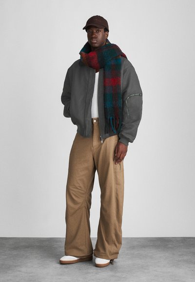 Graue Bomberjacke mit Reißverschluss, weißer Pullover, braune weite Hose und ein mehrfarbiger kariert Schal. Weiße Schuhe runden das Outfit ab.