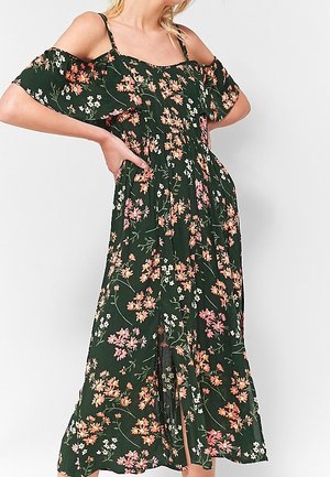 Femme portant une robe maxi verte foncée à fleurs, épaules dénudées, fines bretelles et fente à l’avant, posant les mains sur les hanches.