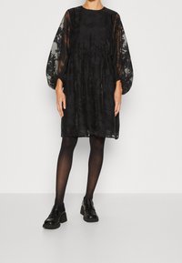 Robe noire avec manches longues transparentes à motifs, coupe décontractée, taille élastique, associée à des chaussures noires et des collants.