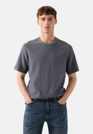 Ung mand med kort brunt hår iført en mørkegrå kortærmet striktrøje og blå jeans, stående med hænderne i lommerne.