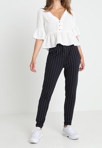 Biała bluzka peplum z rękawami w trzech czwartych i detalami na guziki, w zestawieniu z dopasowanymi spodniami w czarno-białe paski oraz białymi trampkami.