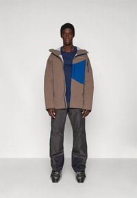 Barna és szürke szigetelt kabát kék díszzsebbel, szürke nadrággal és fekete csizmával párosítva. Kapucnival és cipzáras zsebekkel rendelkezik.