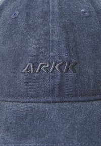 Cappello in denim blu scuro con logo "ARKK" ricamato sul davanti, con cuciture visibili e una visiera strutturata.