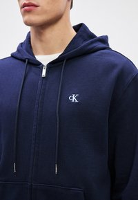 Hanorac cu fermoar de culoare albastru marin, fabricat dintr-un material moale, dispune de un buzunar tip cangur și șnururi cu capete din metal, având un mic logo brodat.