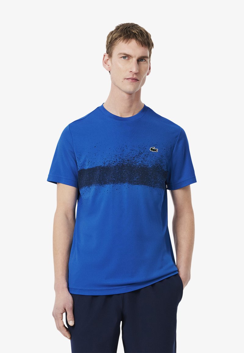 Camiseta azul de manga corta en tela suave con un diseño de salpicaduras negras en degradado en el pecho. Presenta un pequeño logo verde a la izquierda.
