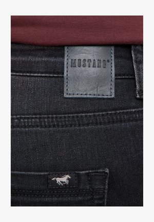 Jean en denim noir avec un patch en cuir texturé portant l'inscription "MUSTANG" et un petit logo de cheval blanc sur la poche arrière.