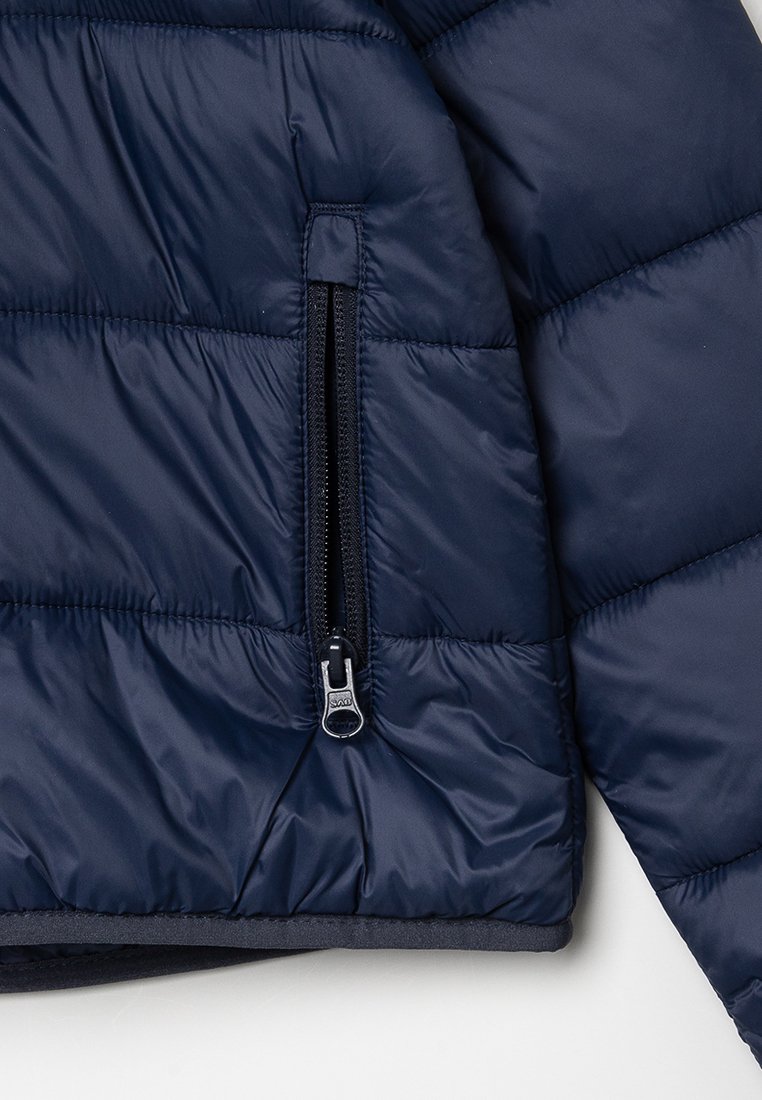 Giacca imbottita blu navy con design a trapunta, dotata di tasca con zip e texture liscia. Dettaglio della zip visibile.
