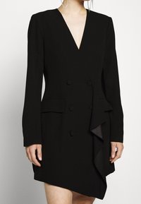 Robe noire avec un décolleté en V profond, manches longues, design croisé à double boutonnage, détail volanté à l'avant, et une ourlet effilé. Confectionnée dans un tissu lisse.