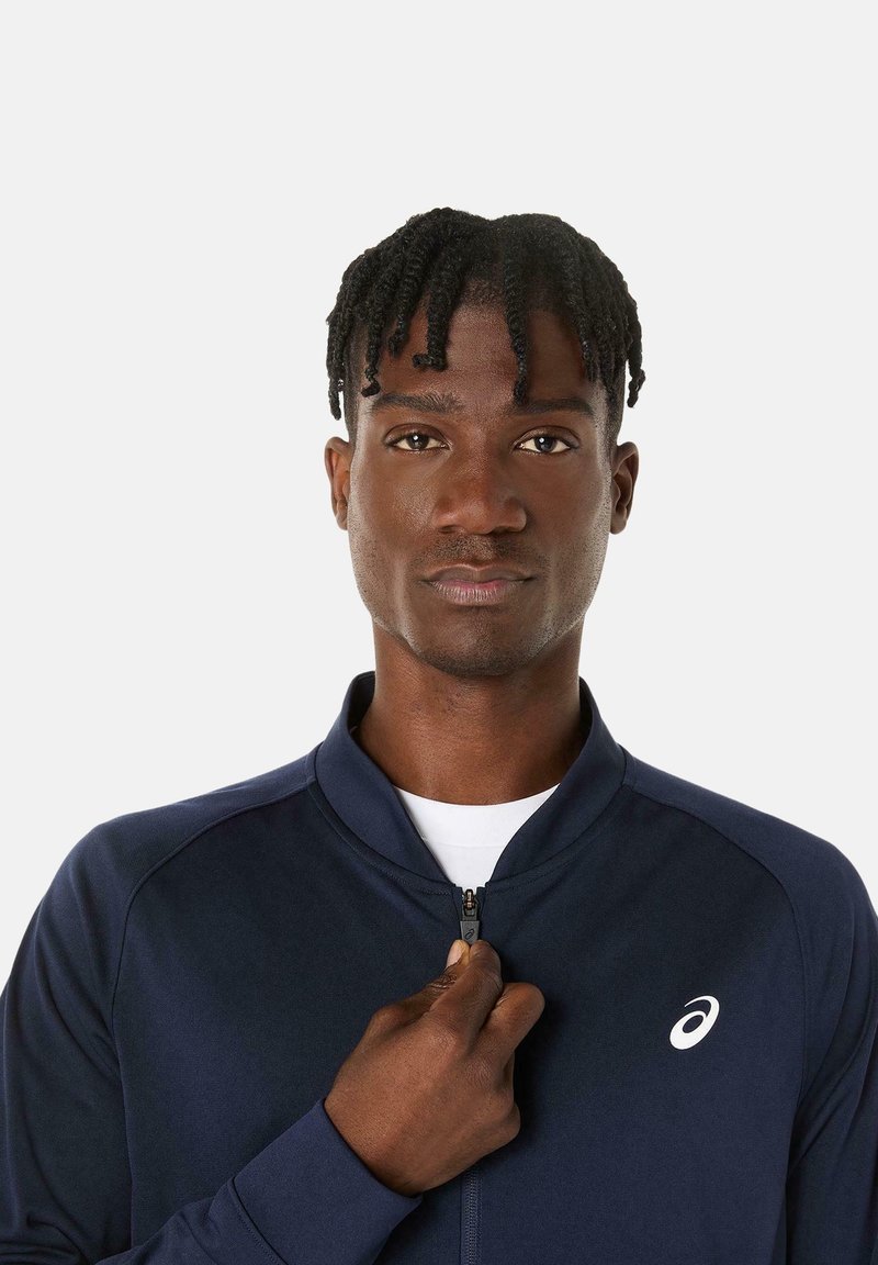 Giacca atletica navy blu con zip e colletto rialzato; presenta il logo bianco sul lato sinistro del petto e una texture liscia.