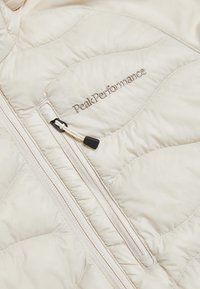 Giacca isolante beige chiaro con texture trapuntata, dotata di una tasca laterale con zip e logo "Peak Performance" ricamato in oro.