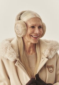 Béžové nausníky s kožušinovou výstelkou a výšivkou medveďa, ktoré nosí žena v béžovom kabáte s kožušinovým golierom a nášivkou, na neutrálom pozadí.