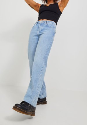 Person wearing light blue straight-leg jeans, black tank top, black boots, posing with arms raised against a plain white background.