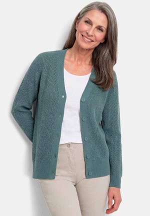 Frau mit langen grauen Haaren lächelt, trägt eine türkisfarbene, geknöpfte Strickjacke über einem weißen Hemd und beigen Hosen und steht vor einem hellen Hintergrund.