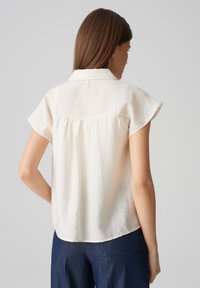 Helles beiges Kurzarmhemd mit Kragen, mit gerafftem Rückendetail und einer zerknitterten Textur, kombiniert mit dunkelblauen Hosen.