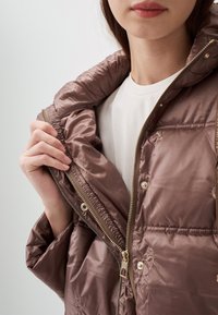 Braune Pufferjacke mit gl�änzender, gesteppten Textur. Verfügt über einen Reißverschluss vorne, elastische Bündchen und dekorative Nähte. Die Innenschicht ist glatt.