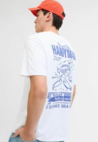 T-shirt blanc à manches courtes avec une impression graphique bleue comportant du texte et un bricoleur en bande dessinée au dos. Matière en coton lisse.