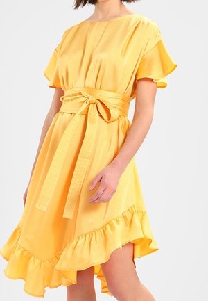 Femme portant une robe jaune vif avec un ourlet à volants, des manches flottantes et une large ceinture nouée à la taille, sur un fond uni.