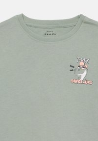 Žalias medvilninis marškinėliai su apvaliu kaklu, puošiami rožiniu ir juodu T-Rex grafiku su akiniais nuo saulės. Glotni tekstūra, laisvas kirpimas.