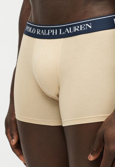 Homme portant un boxer beige Polo Ralph Lauren avec une ceinture bleu marine, debout les bras détendus le long du corps.