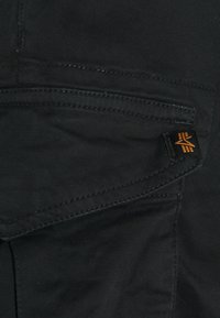 Närbild av en svart tygficka med synliga stygn och en liten svart etikett med en orange geometrisk logotyp.