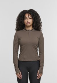 Brown long-sleeve athletic top with a half-zip collar, fitted design, and seam detailing. Paired with black high-waisted leggings.