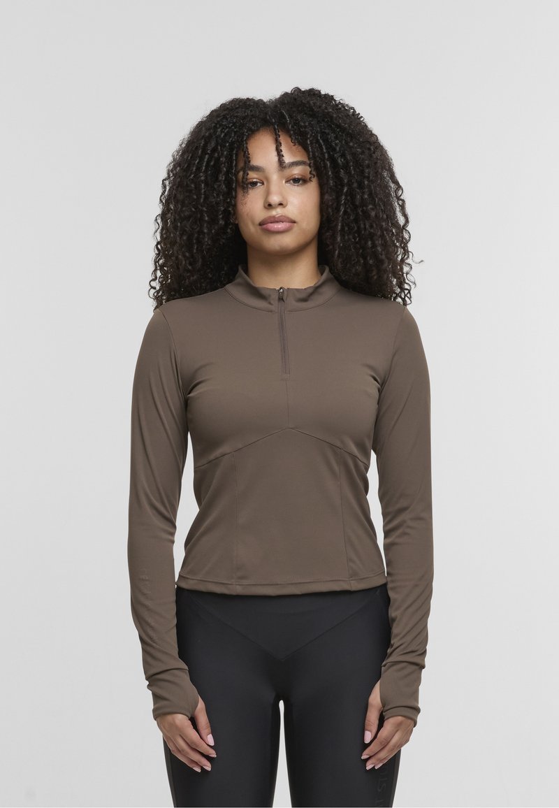 Brown long-sleeve athletic top with a half-zip collar, fitted design, and seam detailing. Paired with black high-waisted leggings.