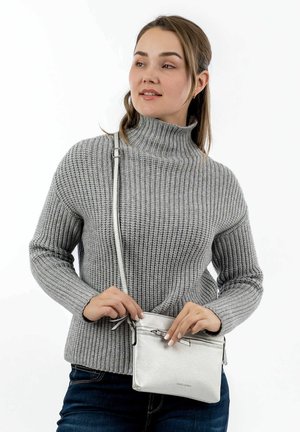Junge Frau in einem grauen, gerippten Rollkragenpullover, die eine kleine silberne Umhängetasche anpasst, vor einem weißen Hintergrund stehend.