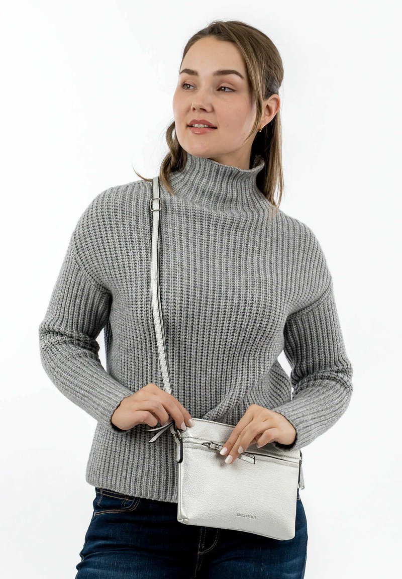 Joven mujer con un suéter de cuello alto gris de canalé ajustando un pequeño bolso cruzado plateado, de pie frente a un fondo blanco.