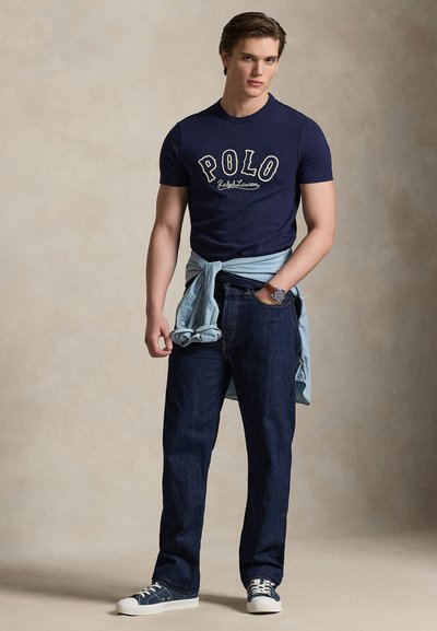 Μπλε ναυτικό βαμβακερό T-shirt με λογότυπο "POLO", σε συνδυασμό με σκούρα τζιν τζιν. Ανοιχτό μπλε πουκάμισο δεμένο γύρω από τη μέση, σε συνδυασμό με λευκά αθλητικά παπούτσια.