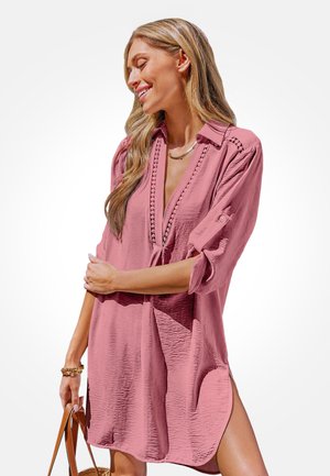 Kobieta uśmiechająca się, ubrana w luźną różową sukienkę z podwiniętymi rękawami i koronkowym wykończeniem, trzymająca brązową torebkę na białym tle.