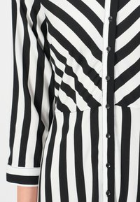 Robe à rayures verticales noires et blanches avec des boutons sur le devant et des manches trois-quarts, gros plan sur le torse et le bras.