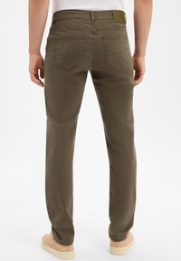 Uomo che indossa pantaloni dritti verde oliva, camicia bianca infilata e scarpe beige, visto da dietro su uno sfondo bianco.