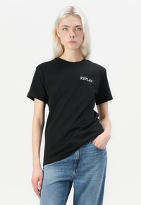 Black cotton t-shirt with short sleeves, featuring a white "REPLAY" logo on the chest. Paired with light blue denim jeans.