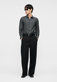 Άνδρας που φοράει σκούρο γκρι πουκάμισο με κουμπιά, μαύρο ψηλόμεσο πλισέ παντελόνι με ζώνη, μαύρα παπούτσια και μαύρα γυαλιά ηλίου, στέκεται με τα χέρια στις τσέπες.