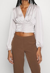 Light gray cropped blouse with v-neck, gathered fabric details, and long puff sleeves. Paired with brown high-waisted trousers.