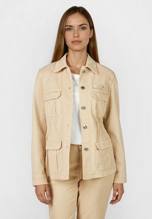 Woman wearing beige buttoned jacket with flap pockets and matching pants, standing against a plain white background.