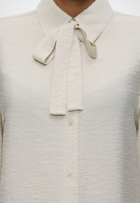 Blouse couleur crème avec un tissu texturé, un col pointu et un nœud noué au cou avec des boutons visibles à l'avant.